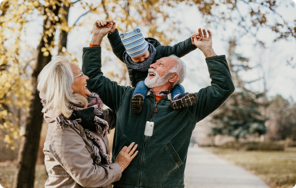 Grandfather wearing a medical alert device holding his grandson up on his shoulders with the grandmother to the left