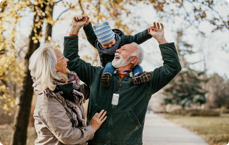 Grandfather wearing a medical alert device holding his grandson up on his shoulders with the grandmother to the left